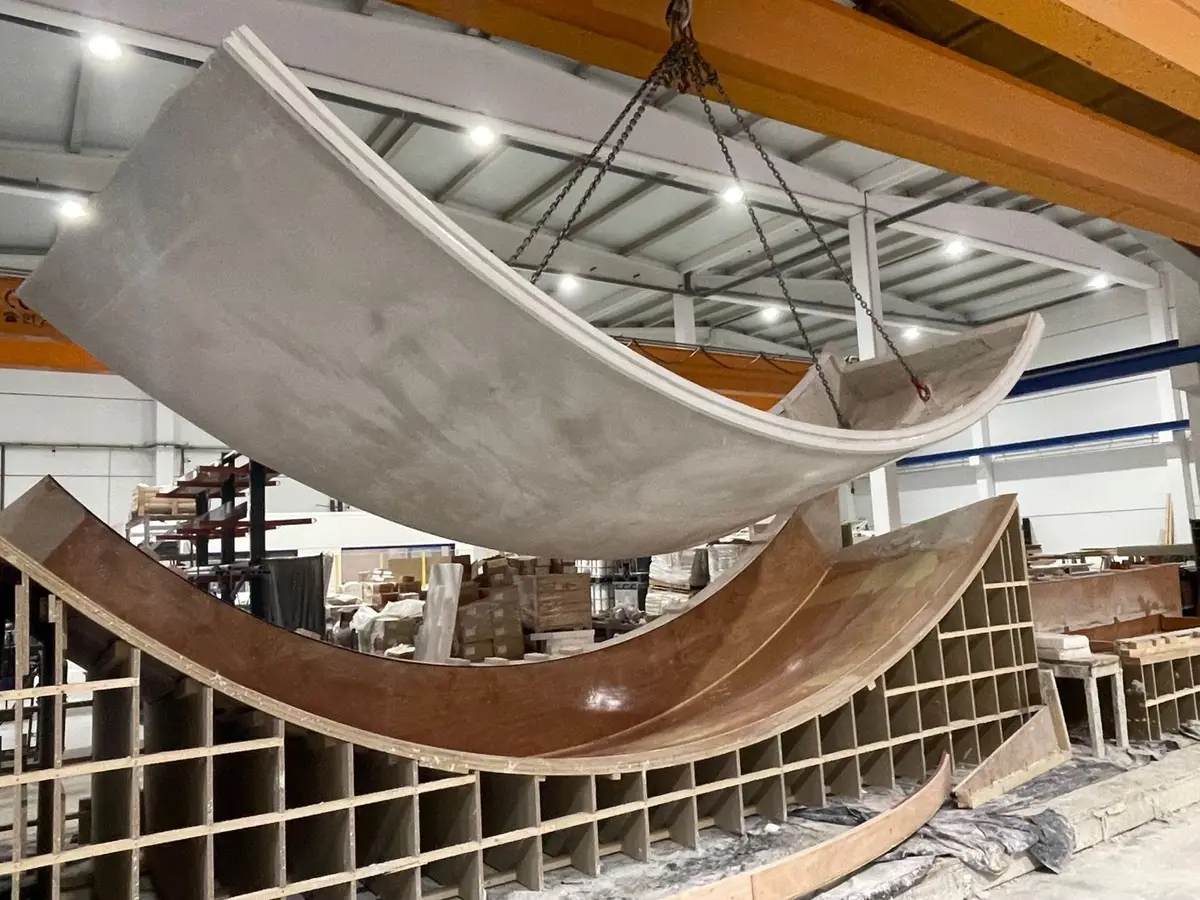 A curved precast shell panel hanging from crane chains above its wooden form inside the plant.