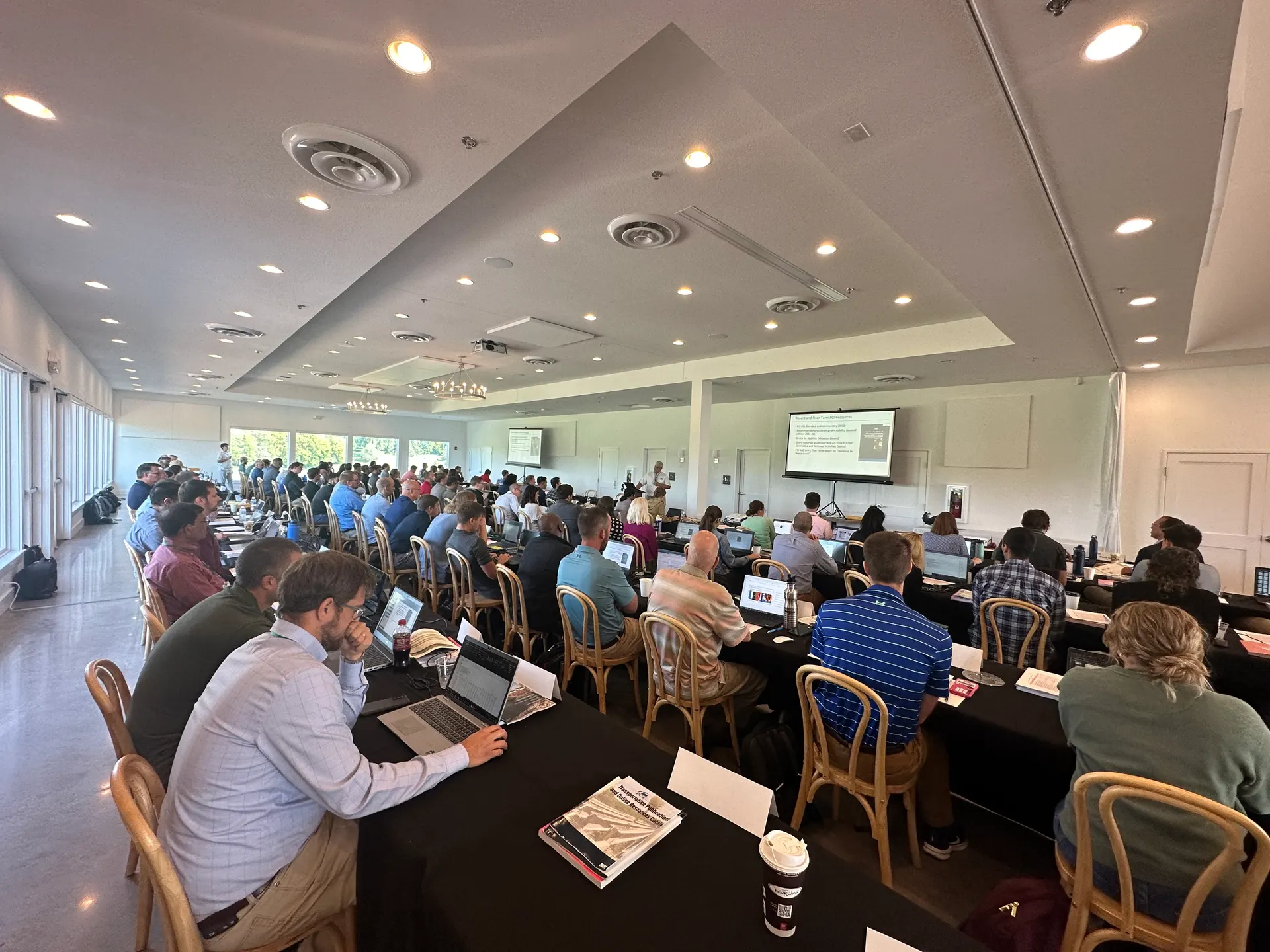 A packed classroom-style seminar with rows of attendees at draped tables facing screens at the far end.