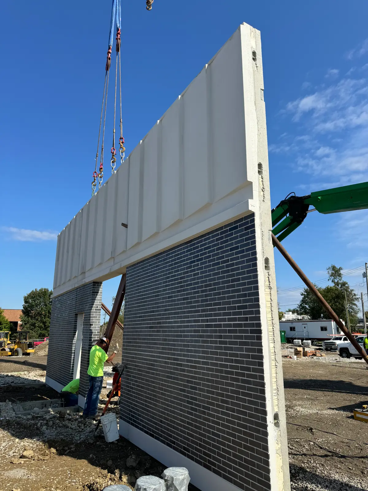 A precast wall panel with dark thin-brick facing being set by crane while a worker steadies its base.