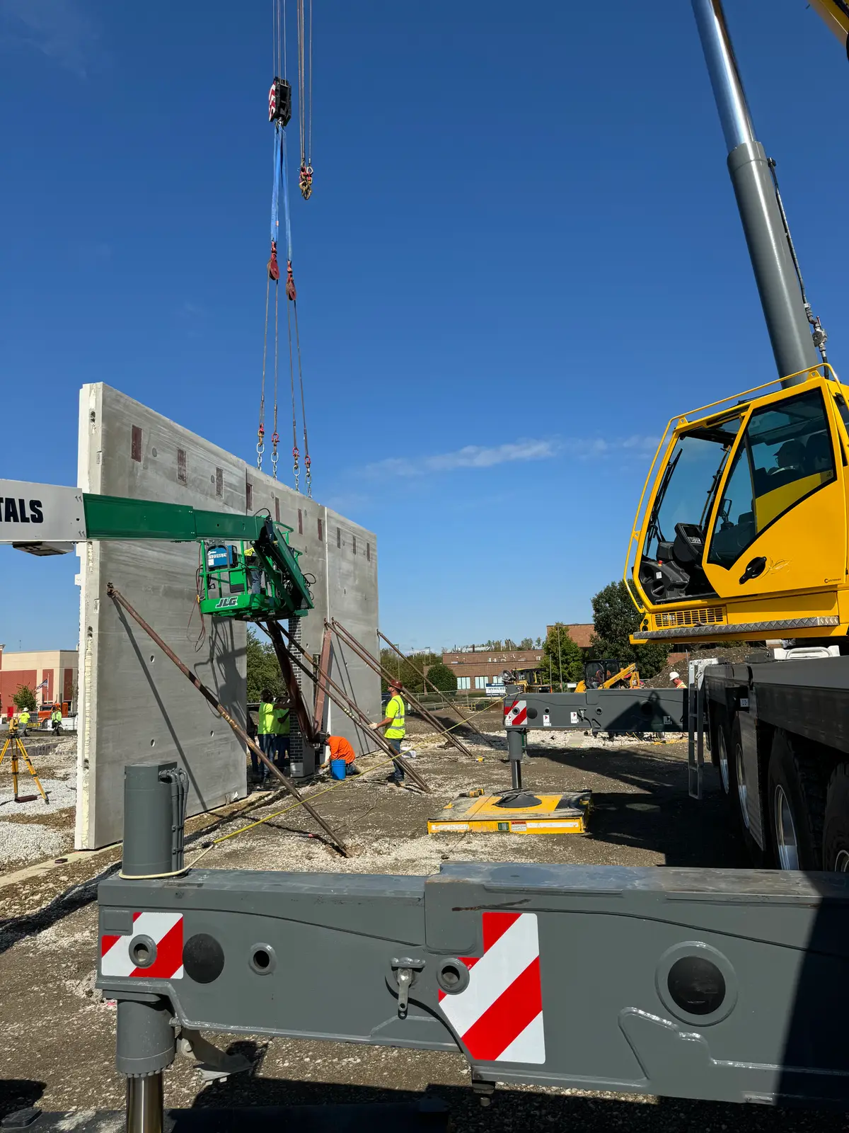 A mobile crane hoisting rigging above a braced concrete wall panel while workers watch from below.