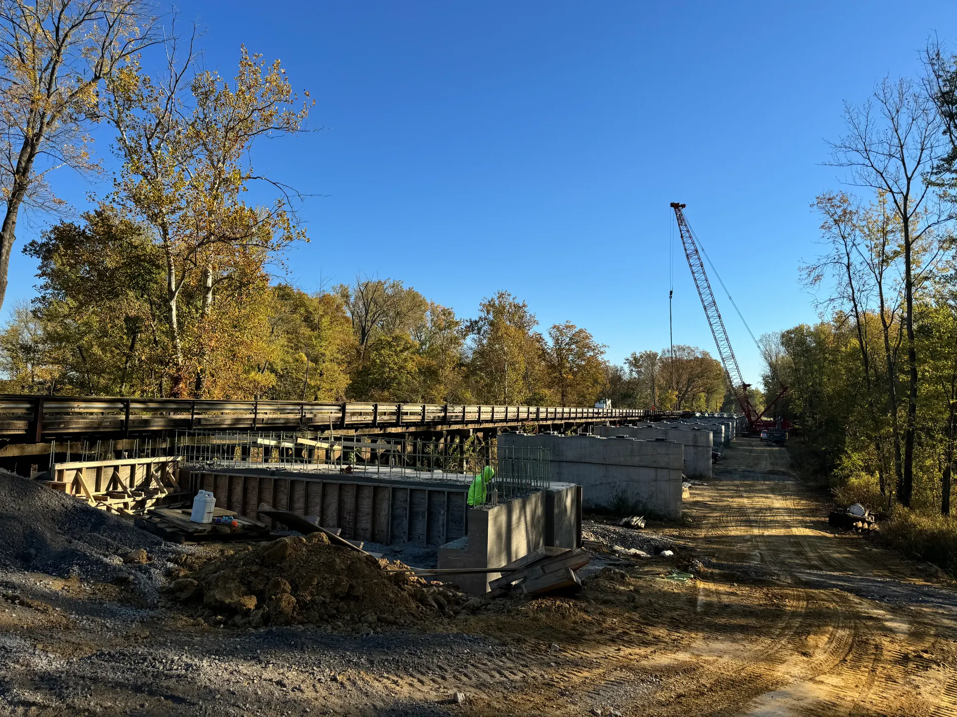 A bridge jobsite in autumn with new concrete abutments in the foreground and a crawler crane down the haul road.