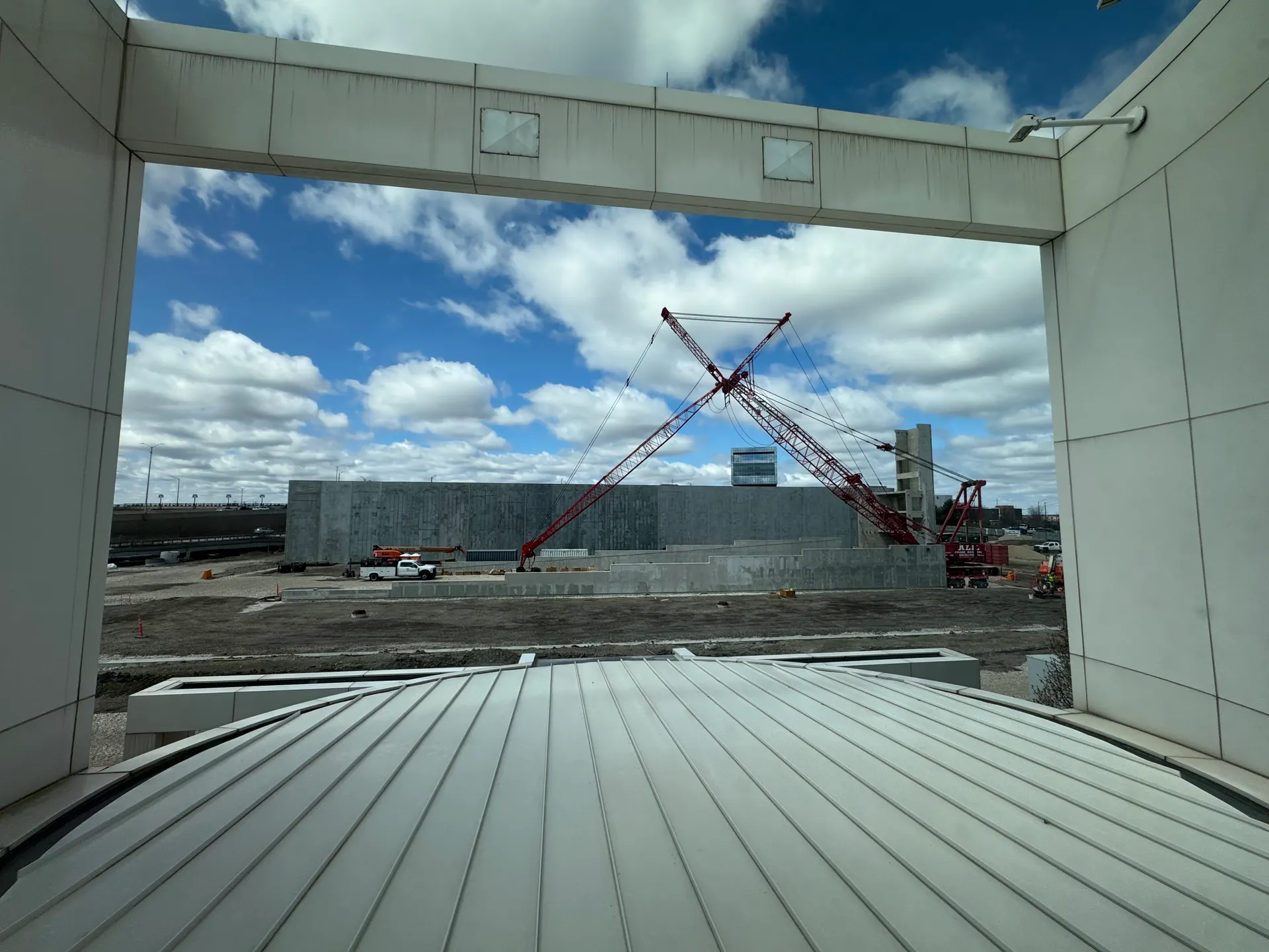 A crawler crane and concrete building shell seen through the frame of a white canopy structure on a jobsite.